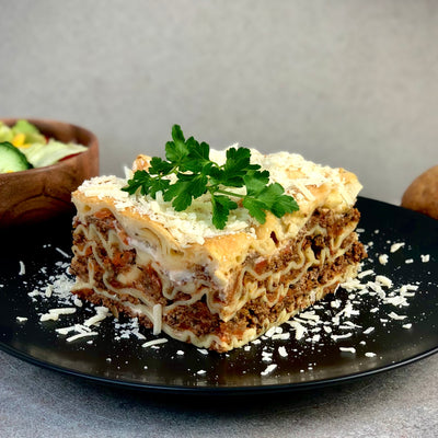 En bit lasagne fotad från sidan där man ser alla lager med köttfärs och lasagneplattor. På sidan finns bröd och en skål sallad som dekoration.