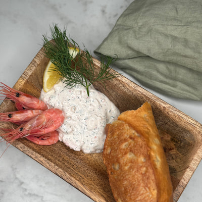 Skagenröra upplagt på ett träfat för inspiration. Pyntat med färska räkor, baguette, dill och citron.